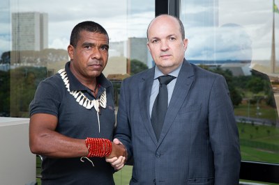 A foto mostra dois homens sérios, apertando as mãos. Um deles é um procurador de terno; o outro, liderança indígena utilizando colares e ornamentos típicos