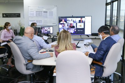 Foto mostra os participantes da videoconferência, sentados em volta de uma mesa retangular.