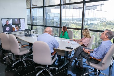 Foto mostra os participantes da videoconferência sentados em volta de uma mesa retangular. à frente, a tv onde os demais participantes aparecem.