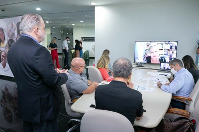 Foto mostra pessoas sentadas à mesa durante reunião, que conta, ainda, com participação de uma mulher por videoconferência