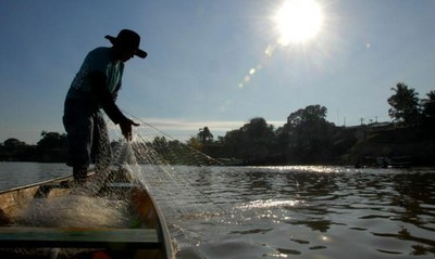 #pracegover: foto de um pescador em pé, em cima de um barco, no momento em que joga a rede de pesca sobre o rio