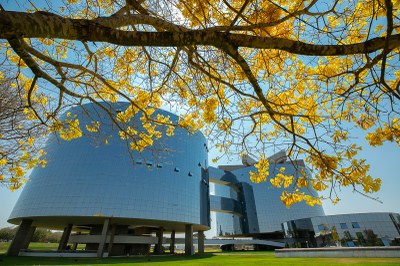 Foto dos prédios que abrigam a procuradoria-geral da república, em brasília. os prédios são redondos, revestidos de vidro e interligados. à frente das edificações há ipês amarelo floridos.