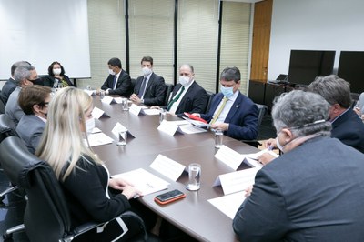 Foto mostra os participantes da reunião sentados em volta de uma grande mesa retangular. Todos estão usando máscara. 