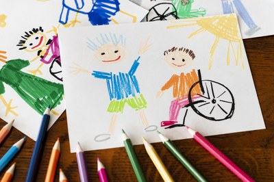 #pracegover: foto registra, sobre a mesa, vários lápis de cores diferentes e desenhos coloridos em que crianças típicas convivem com crianças com deficiência. 
