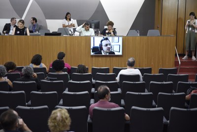Foto do auditório da Assembleia Legislativa de MG com pessoas sentadas nas cadeiras, mesa de debatedores ao fundo e uma tela de videoconferência com a imagem de um homem com fones de ouvido durante sua exposição
