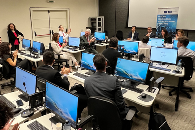 Imagem da sala de aula do curso presencial com os participantes em computadores e uma mesa na frente em que aparecem os palestrantes sentados. 