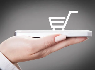 Foto da mão de uma mulher segurando um celular. Sobre o celular, existe a figura de uma carrinho de compras, na cor branca. 
