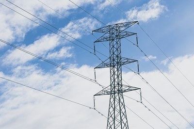 Descrição da imagem #PraTodosVerem: fotografia de torre elétrica, ao fundo, céu azul com nuvens brancas
