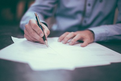 Foto com destaque de mãos segurando caneta e apoiando papel sobre mesa, assinando documento.