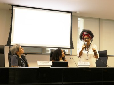 Fotografia de momento do evento, com as palestrantes KK Verdade à esquerda e Vilma Pereira, segurando o microfone à direita. Ao centro, Tatiane Pereira, moderadora e coorganizadora