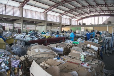 Imagem mostra galpão com várias caixas de papelão com material reciclável e pessoas trabalhando na separação desse material.