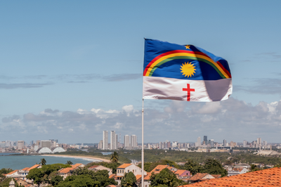 A imagem mostra a bandeira de Pernambuco em primeiro plano e a cidade do Recife ao fundo. 