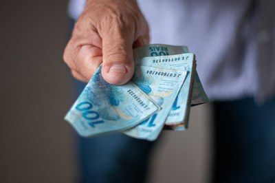 #Pracegover Imagem de um homem (sem mostrar o rosto) usando camisa azul, com a mão estendida, segurando cédulas de 100 reais.