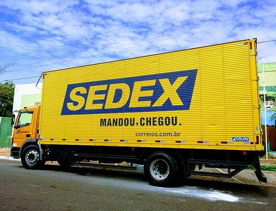 Caminhão de entrega de Sedex estacionado na rua, cabine e caçamba amarelas. Na caçamba, a logo do Sedex e o slogan "Mandou, chegou"