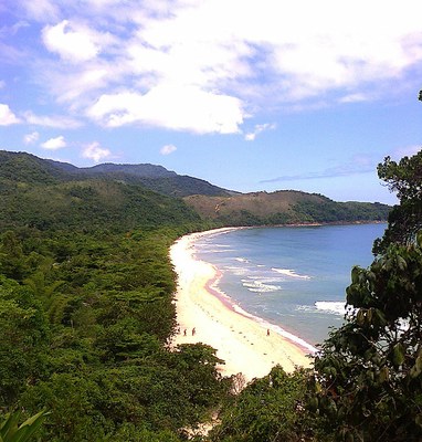 Vista da chegada por trilha na Praia do Sono, com vegetação à esquerda, algumas montanhas ao fundo, faixa de praia com quatro pessoas ao centro e mar à direita. Céu azul com algumas nuvens.