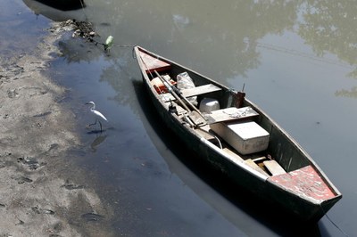 #pracegover: Foto de um barco de pesca parado na beira da Baía de Guanabara, que está poluída. Na foto também há uma garça andando na beira da água. 
