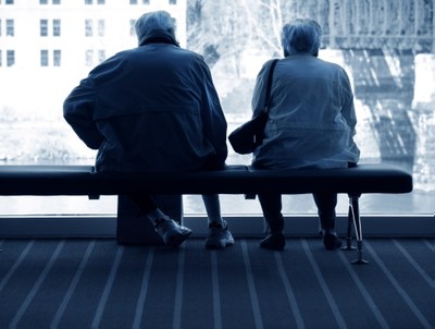Foto em preto e branco com dois idosos sentados de costas num banco no centro da imagem