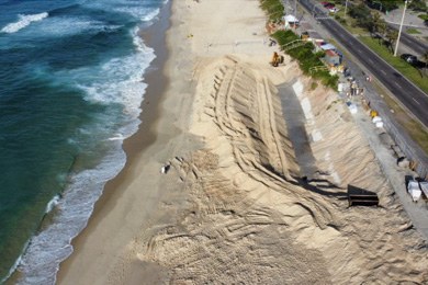 Foto mostra uma faixa de areia no centro, com várias falhas decorrentes de obras. À esquerda se vê o mar e à direita uma avenida com área verde.