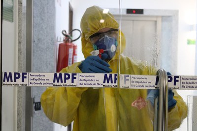 Imagem de pessoa com traje de proteção fazendo limpeza de porta de vidro da PR/RN. 