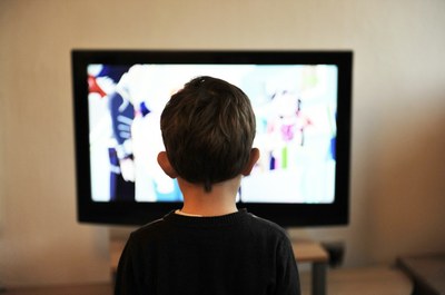 Foto de um menino de costas, não identificável, olhando para um aparelho de tv, no qual também não se pode identificar o conteúdo 