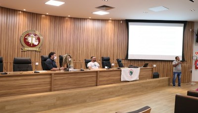 A fotografia retrata quatro pessoas em no auditório do Ministério Público Estadual em Rondônia. Três estão na bancada do auditório e uma quarta pessoal está em pé, de frente para uma tela para projeção. 