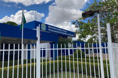 Foto da fachada da sede do Ministério Público Federal em Roraima, um prédio azul e branco de dois andares com o letreiro na parte superior. Uma cerca branca está em primeiro plano.