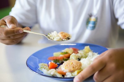 Foto de um prato de comida em primeiro plano e uma criança segurando uma colher cheia de comida em direção à boca
