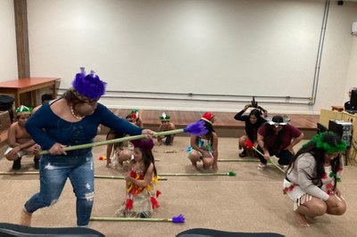 Foto mostra cerca de dez pessoas indígenas realizando uma espécie de dança, com enfeites e objetos típicos, numa sala de paredes brancas e chão bege.