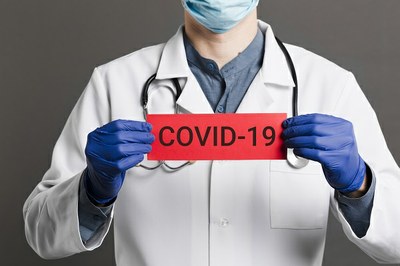 #Pracegover Foto de profissional de saúde com jaleco branco, luvas azuis e máscaras, segurando uma placa vermelha onde está escrito covid-19
