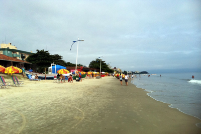 Praia com faixa de areia à esquerda e mar à direita. Há imóveis construídos na faixa de areia, além de guarda-sóis e pessoas caminhando na praia 
