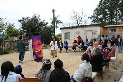 Ao fundo, numa área externa, uma construção em alvenaria que faz parte da comunidade Toca Santa Cruz. Mais à frente, diversas pessoas sentadas, formando um semicírculo, acompanham a fala de uma mulher em pé. À esquerda, uma artista grafita uma geladeira, que exibe a ilustração de uma mulher sorrindo.
