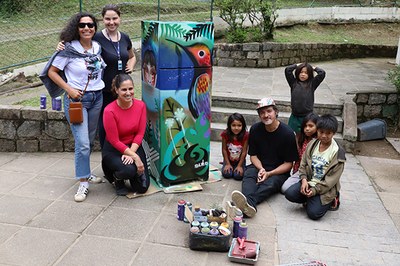 Uma geladeira grafitada com o desenho de um beija-flor. À esquerda, três servidoras do MPF. À direita, o pintor Ricca de Lucca e quatro crianças indígenas.