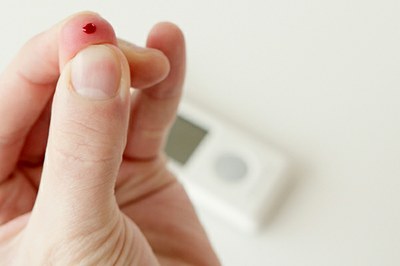 Em primeiro plano, uma gota de sangue na ponta do dedo de uma mão. Ao fundo, um glicosímetro.