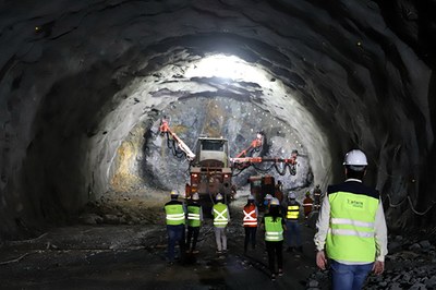 Dentro de um túnel em construção, se vê, em primeiro plano, seis pessoas com capacetes e coletes de proteção. Ao fundo, uma máquina trabalha na escavação.