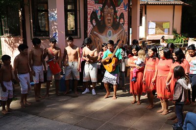 Ao centro, em pé, um menino indígena toca um violão. À esquerda, outros seis meninos em pé, lado a lado, observam o violonista. À direita, também lado a lado, quatro meninas indígenas acompanham a apresentação.
