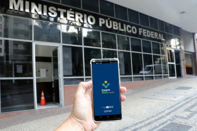 Mão de uma pessoa segurando celular mostrando a tela inicial do aplicativo Conect SUS. Ao fundo, prédio com nome Ministério Público Federal.