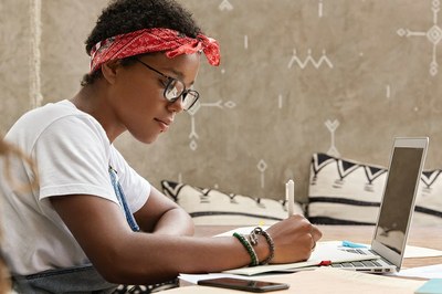 Foto de estudante negra sentada à mesa escrevendo em um papel em frente a um notebook