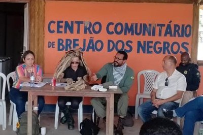 Foto de cinco pessoas sentadas à mesa. Na parede de fundo, de cor laranja, lê-se: Centro Comunitário de Brejão dos Negros. 