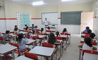 Crianças assistem aula sentadas, diante da professora. 