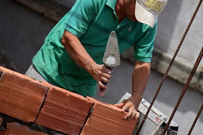 #Paratodosverem: Imagem mostra trabalhador de camisa polo verde claro e boné cor creme claro assentando um bloco cerâmico com uma pá de pedreiro em uma parede que está parcialmente construída até meia altura.