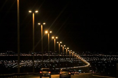 #PraTodosVerem: foto colorida de uma estrada com automóveis à noite, com os postes de luz acesos e, ao fundo, as luzes de uma cidade.