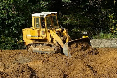 #Paratodosverem: Imagem mostra escavadeira de pequeno porte movendo terra sobre um terreno irregular. Um homem de jaqueta preta a dirige. Ao fundo, um trecho de mata