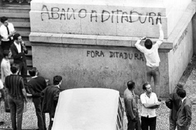 #PraCegoVer: Foto de época mostra homem pixando a frase "Abaixo a Ditadura" em um muro