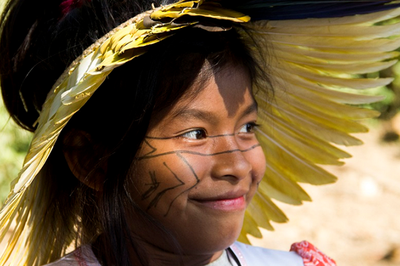 Foto de uma criança indígena com rosto pintado e cocar. Ela olha para o lado e sorri 