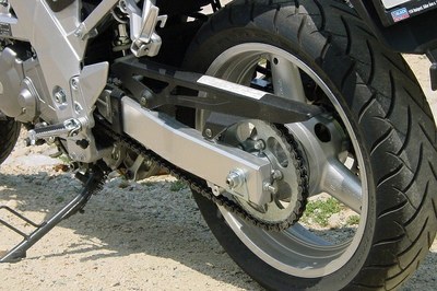 #Paratodosverem: Imagem mostra detalhe de uma motocicleta cinza estacionada sobre um solo seco, com destaque para a roda traseira e parte do sistema de transmissão