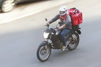#Paratodosverem: imagem mostra homem guiando uma motocicleta. Ele está com uma jaqueta cinza, calça jeans azul, capacete branco e carrega nas costas uma mochila em formato de cubo vermelha, típica de entregadores por aplicativo