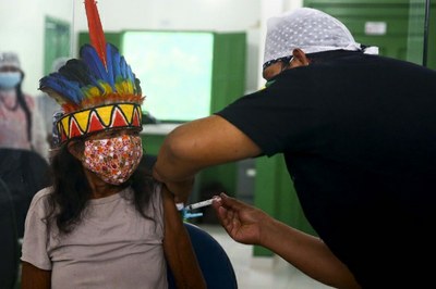 #pracegover: imagem colorida mostra um agente de saúde vacinando uma indígena com cocar, ambos usando máscara.