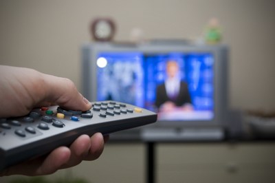Mão segurando controle remoto, com aparelho de TV ao fundo. 